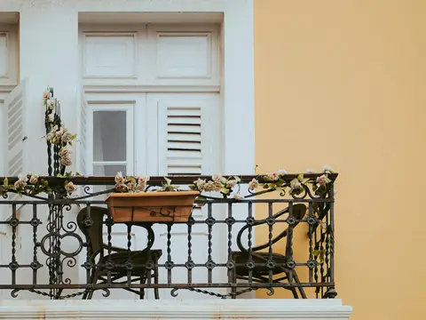 Balcon d'une façade jaune et blanche fermé par un garde-corps de fer noir à barreaux torsadés et rosaces, avec une jardinière fleurie et deux chaises