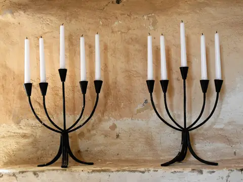 Deux candélabres en fer forgé noir à cinq branches portant des bougies blanches, posés sur une tablette devant un mur en enduit ancien