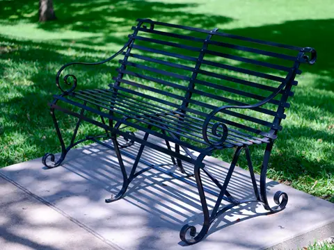 Banc en fer forgé noir à lames, accoudoirs à volutes et pieds en spirale, posé sur une dalle de béton devant une pelouse ensoleillée