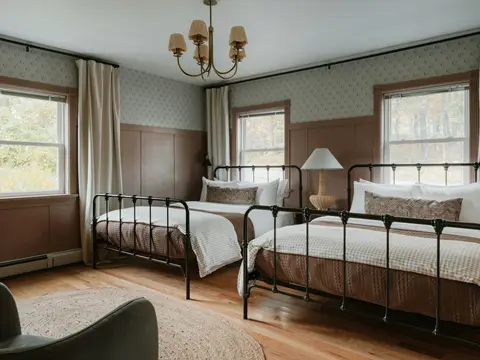 Chambre de charme à lambris et papier peint : deux lits en fer forgé noir à barreaux, lustre à bras de laiton, parquet clair et tapis tressé