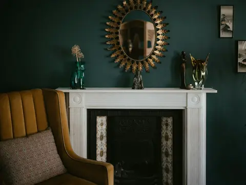 Miroir ovale entouré de feuilles de laiton doré au-dessus d'une cheminée en marbre blanc, mur vert profond et fauteuil velours moutarde