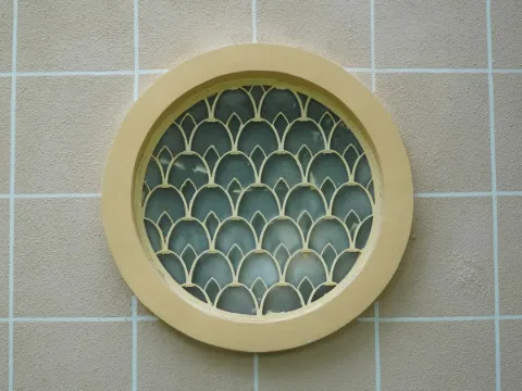 Petite fenêtre ronde au cadre crème, fermée par une grille ajourée à motif d'écailles