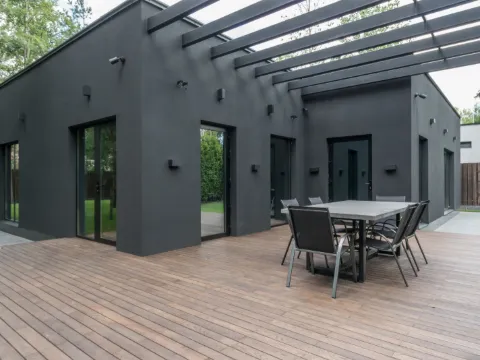 Poutres noires d'une pergola au-dessus d'une terrasse en bois et d'une table, devant une villa noir mat