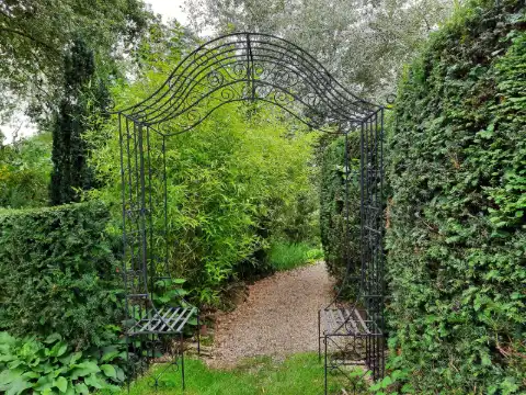 Arche en fer forgé noir à volutes avec assises intégrées, ouvrant sur une allée de gravier entre deux haies